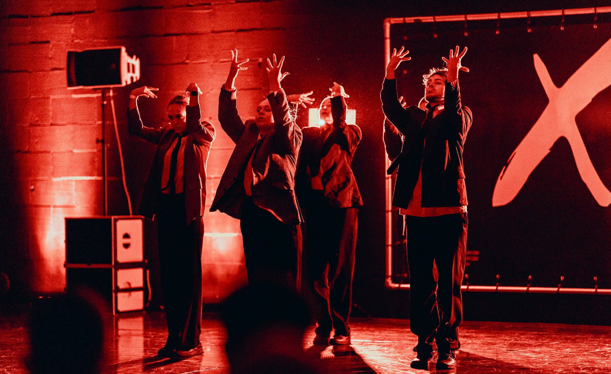 Group of people on a stage with raised hands, red lighting, and a large 'X' logo in the background.